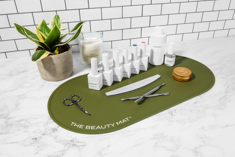 An olive green oval beauty mat labeled 'THE BEAUTY MAT' sits on a white marble countertop, surrounded by nail care products, tools, and a small potted plant against a white subway tile wall.