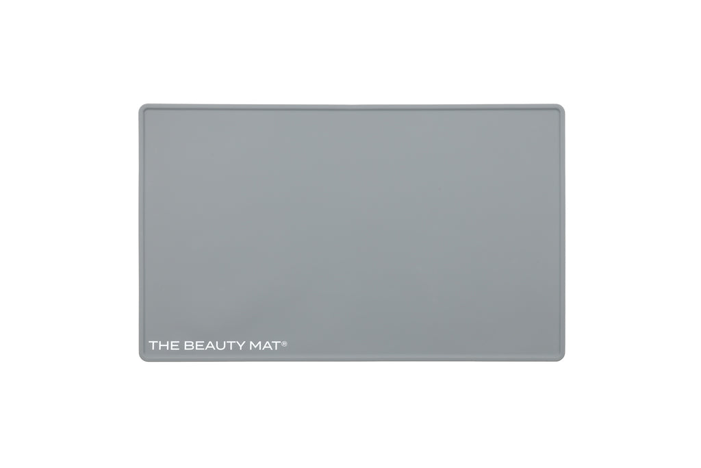 A rectangular ash gray beauty mat with rounded corners and the text 'THE BEAUTY MAT' printed in white at the bottom left, shown on a plain white background.