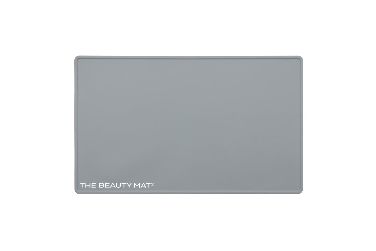 A rectangular ash gray beauty mat with rounded corners and the text 'THE BEAUTY MAT' printed in white at the bottom left, shown on a plain white background.