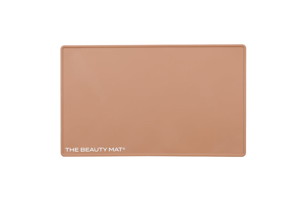 A rectangular caramel beige beauty mat, featuring the text 'THE BEAUTY MAT' printed in white at the bottom left, set against a plain white background.