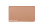 A rectangular caramel beige beauty mat, featuring the text 'THE BEAUTY MAT' printed in white at the bottom left, set against a plain white background.