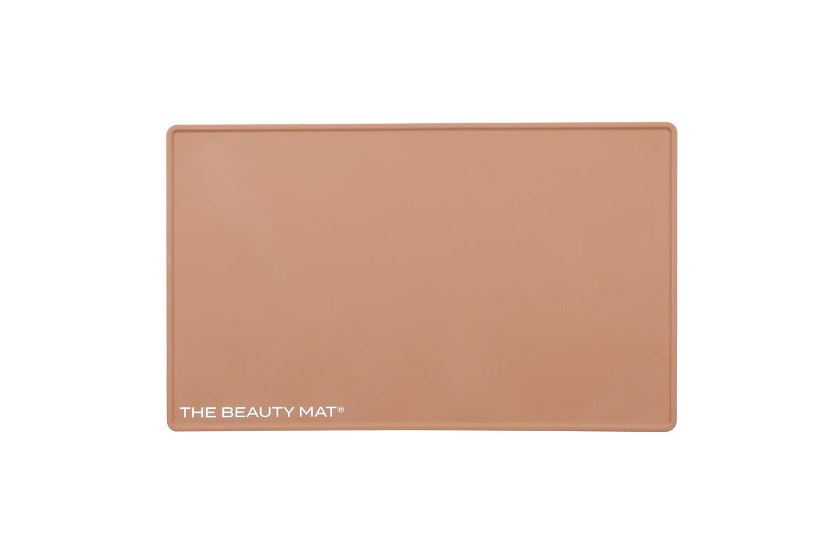 A rectangular caramel beige beauty mat, featuring the text 'THE BEAUTY MAT' printed in white at the bottom left, set against a plain white background.