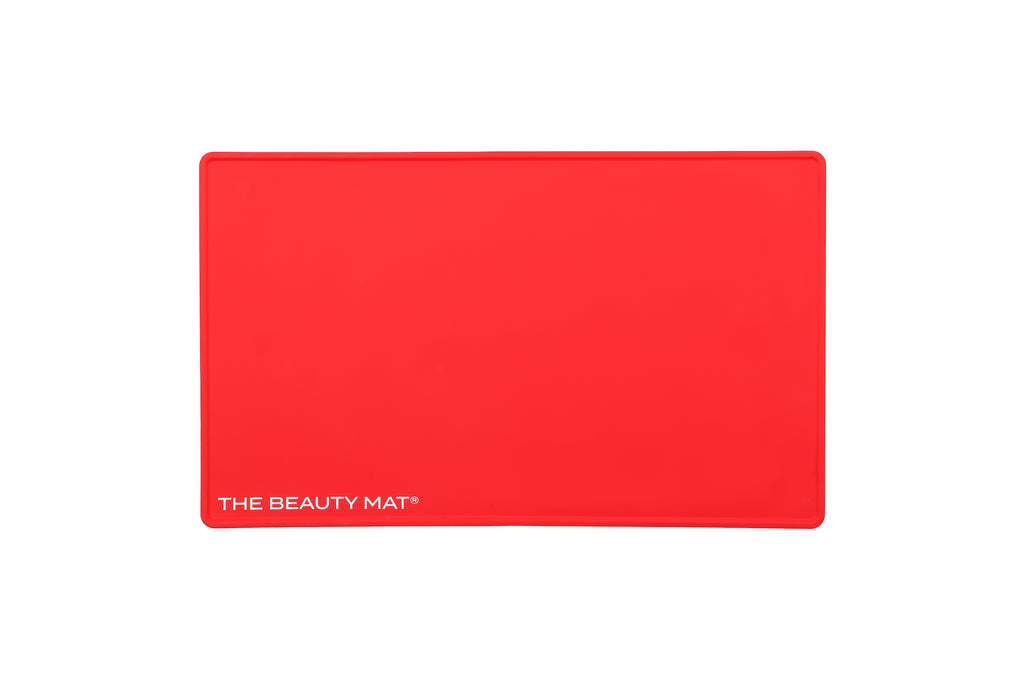 A bright cherry red rectangular silicone beauty mat with rounded corners and the white text 'THE BEAUTY MAT' printed in the bottom-left corner, shown on a plain white background.