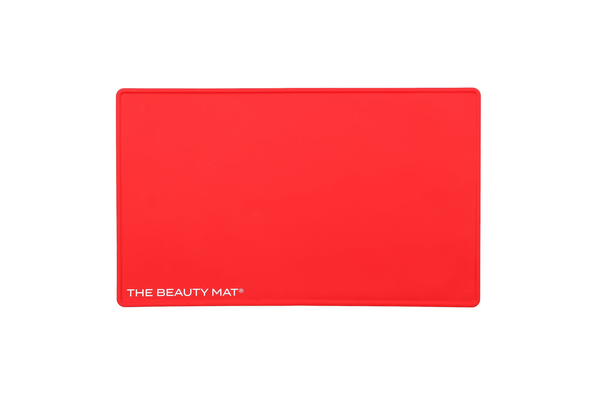 A bright cherry red rectangular silicone beauty mat with rounded corners and the white text 'THE BEAUTY MAT' printed in the bottom-left corner, shown on a plain white background.