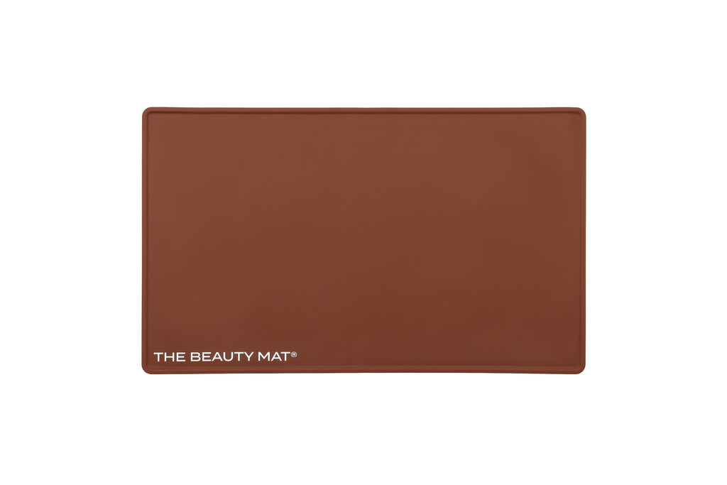 A rectangular chocolate brown silicone beauty mat with rounded corners and the white text 'THE BEAUTY MAT' printed in the bottom-left corner, shown against a plain white background.