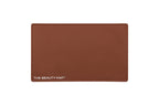 A rectangular chocolate brown silicone beauty mat with rounded corners and the white text 'THE BEAUTY MAT' printed in the bottom-left corner, shown against a plain white background.