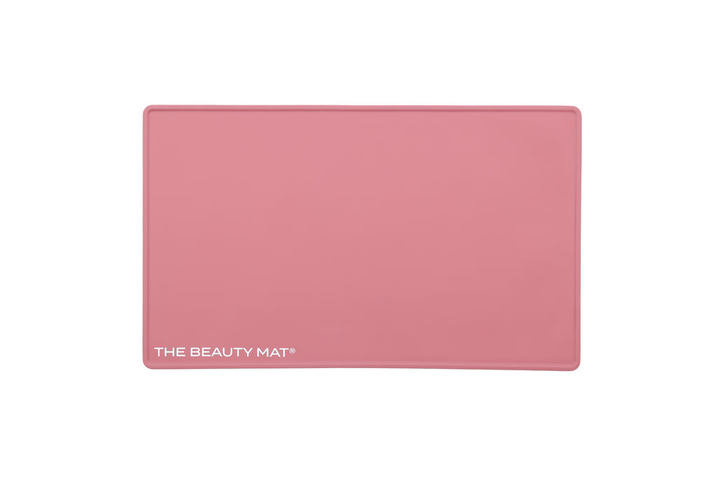 A rectangular dusky pink beauty mat with rounded corners and the text 'THE BEAUTY MAT' printed in white at the bottom left, shown on a plain white background.