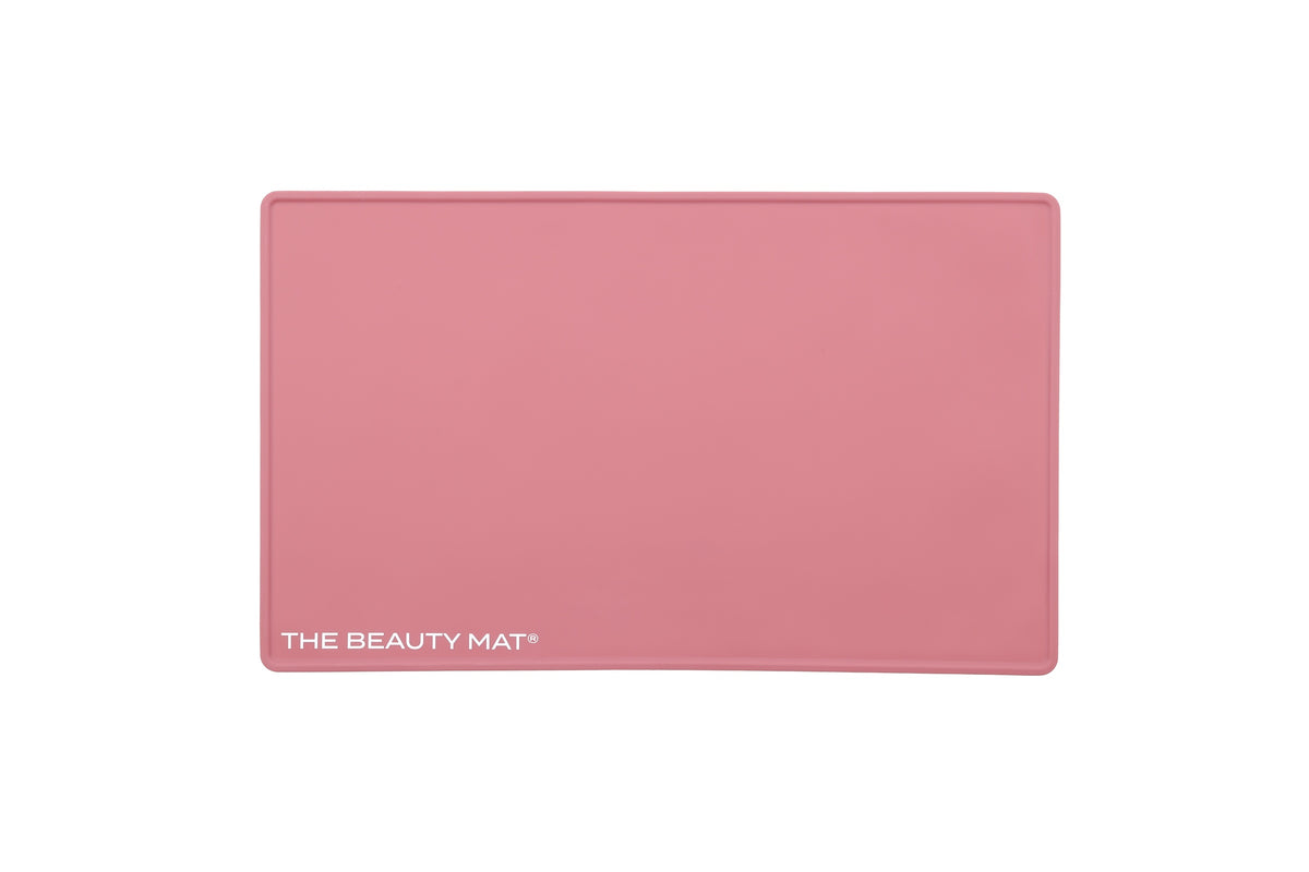 A rectangular dusky pink beauty mat with rounded corners and the text 'THE BEAUTY MAT' printed in white at the bottom left, shown on a plain white background.