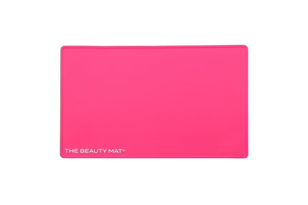 A hot pink rectangular beauty mat with rounded corners and the white text 'THE BEAUTY MAT' printed in the bottom-left corner, shown on a plain white background.