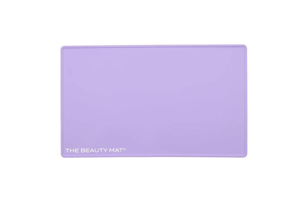 A lilac purple rectangular silicone beauty mat with rounded corners and the text 'THE BEAUTY MAT' printed in white in the bottom-left corner, shown on a solid white background.