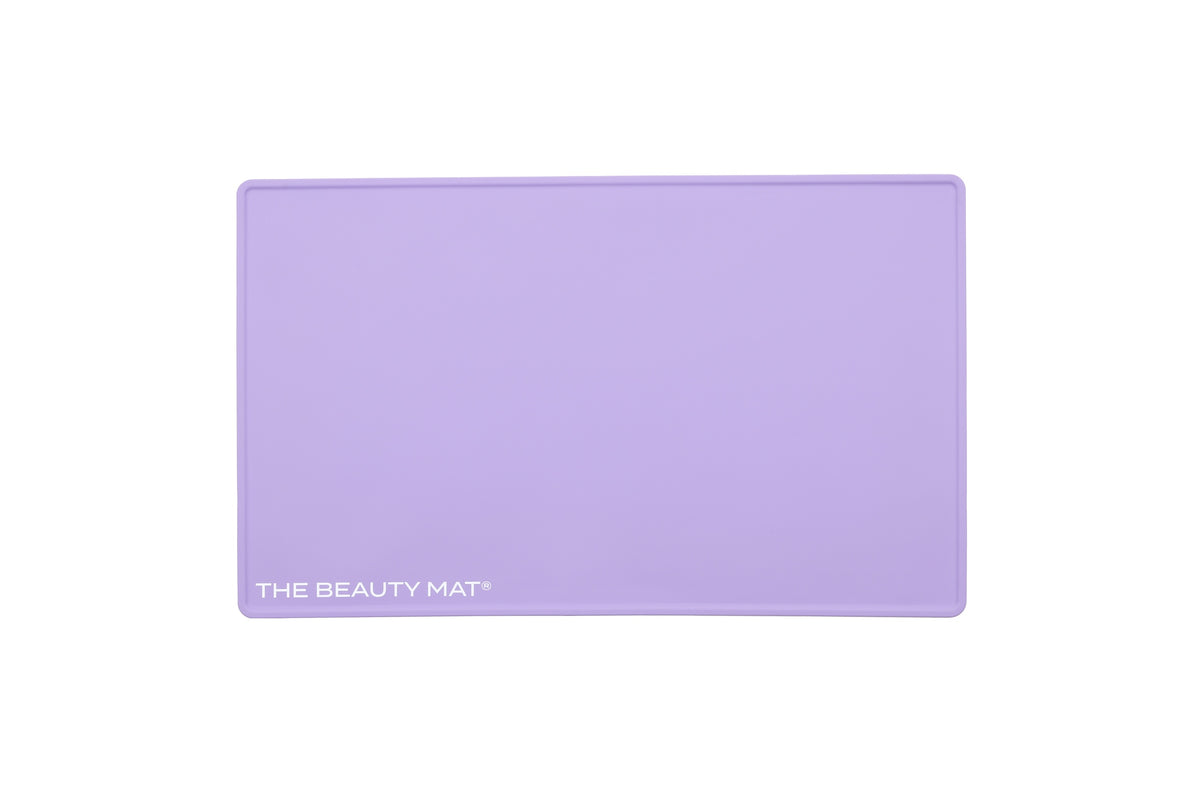 A lilac purple rectangular silicone beauty mat with rounded corners and the text 'THE BEAUTY MAT' printed in white in the bottom-left corner, shown on a solid white background.