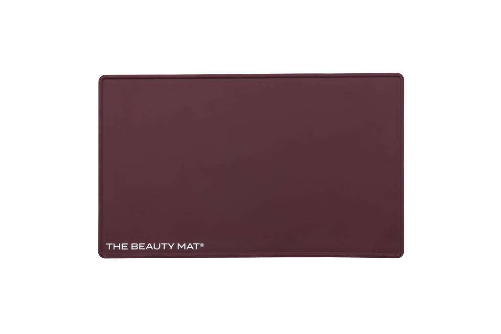 A rectangular maroon beauty mat with rounded corners and the text 'THE BEAUTY MAT' printed in white at the bottom left, shown on a plain white background.