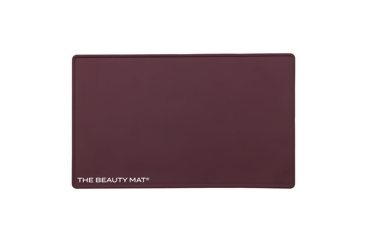 A rectangular maroon beauty mat with rounded corners and the text 'THE BEAUTY MAT' printed in white at the bottom left, shown on a plain white background.
