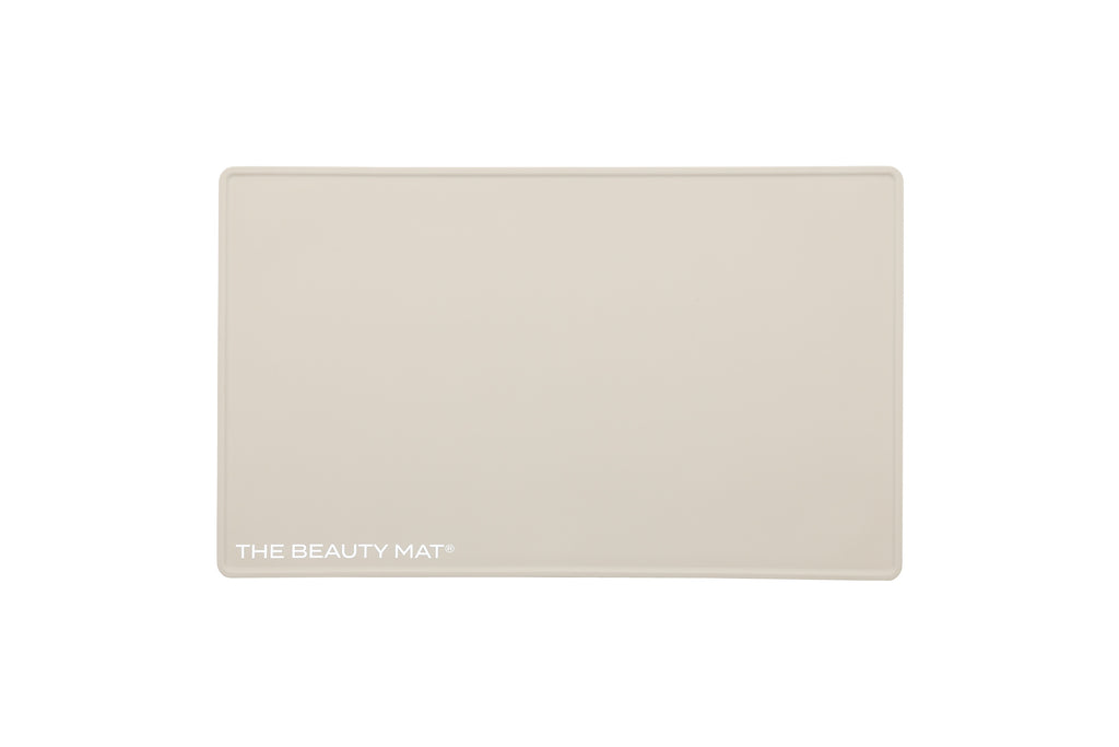 A rectangular oatmeal beige beauty mat with rounded corners and the text 'THE BEAUTY MAT' printed in white at the bottom left, shown on a solid white background.