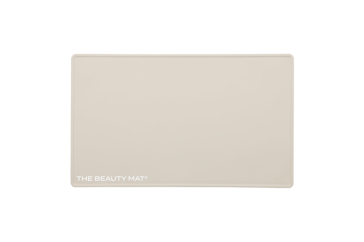 A rectangular oatmeal beige beauty mat with rounded corners and the text 'THE BEAUTY MAT' printed in white at the bottom left, shown on a solid white background.