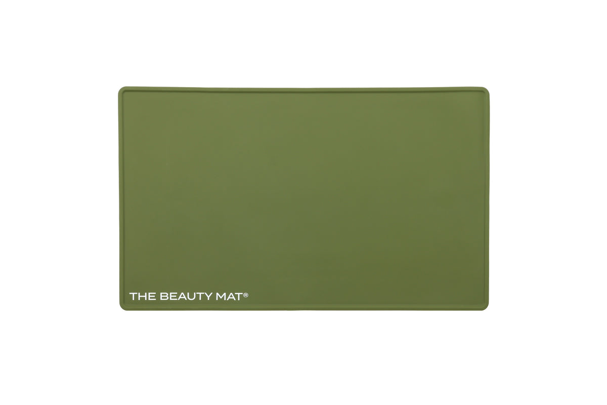 An olive green rectangular silicone beauty mat with rounded corners, featuring the white text 'THE BEAUTY MAT' in the bottom-left corner, shown against a plain white background.