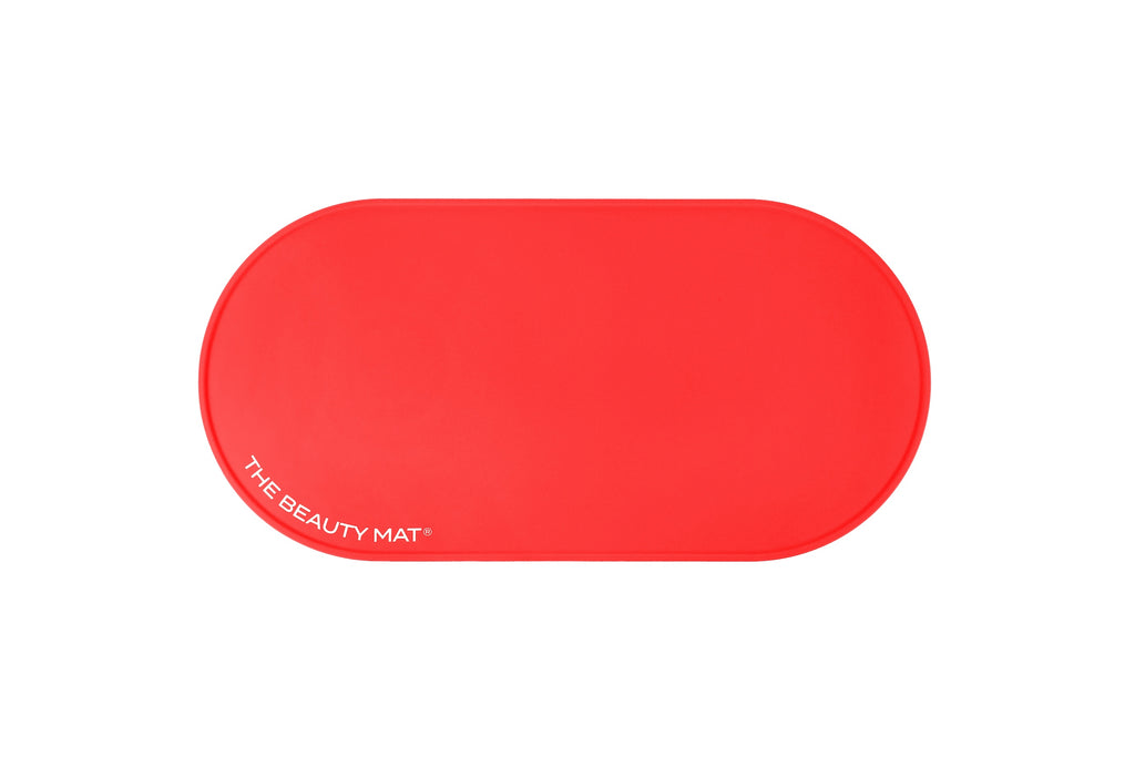 A bright cherry red, oval-shaped silicone beauty mat with rounded corners, featuring the white text 'THE BEAUTY MAT' printed on the bottom-left edge, set against a plain white background.
