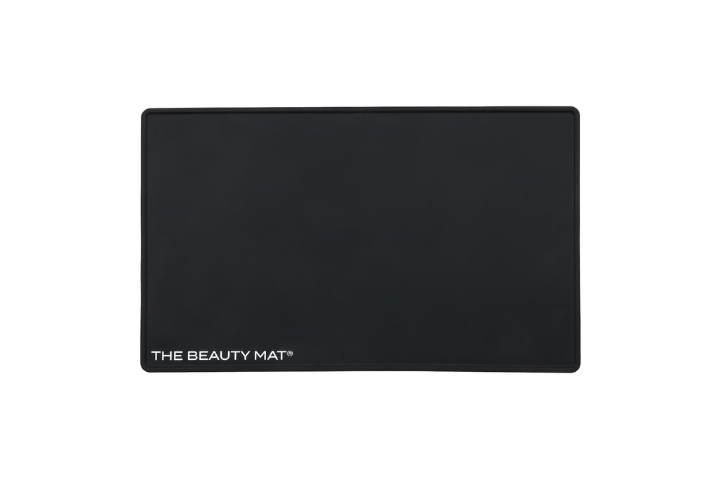 A black rectangular beauty mat with rounded corners and the text 'THE BEAUTY MAT' printed in white at the bottom left, shown on a plain white background.
