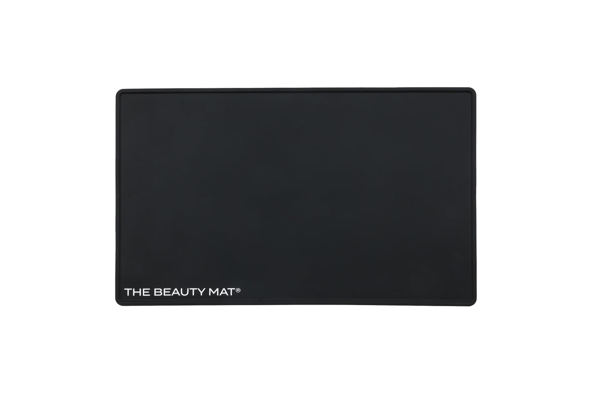 A black rectangular beauty mat with rounded corners and the text 'THE BEAUTY MAT' printed in white at the bottom left, shown on a plain white background.