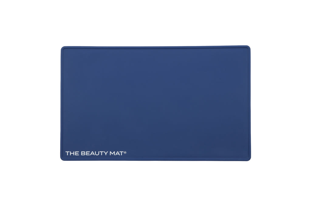 A rectangular navy blue beauty mat with rounded corners and the text 'THE BEAUTY MAT' printed in white at the bottom left, shown on a plain white background.