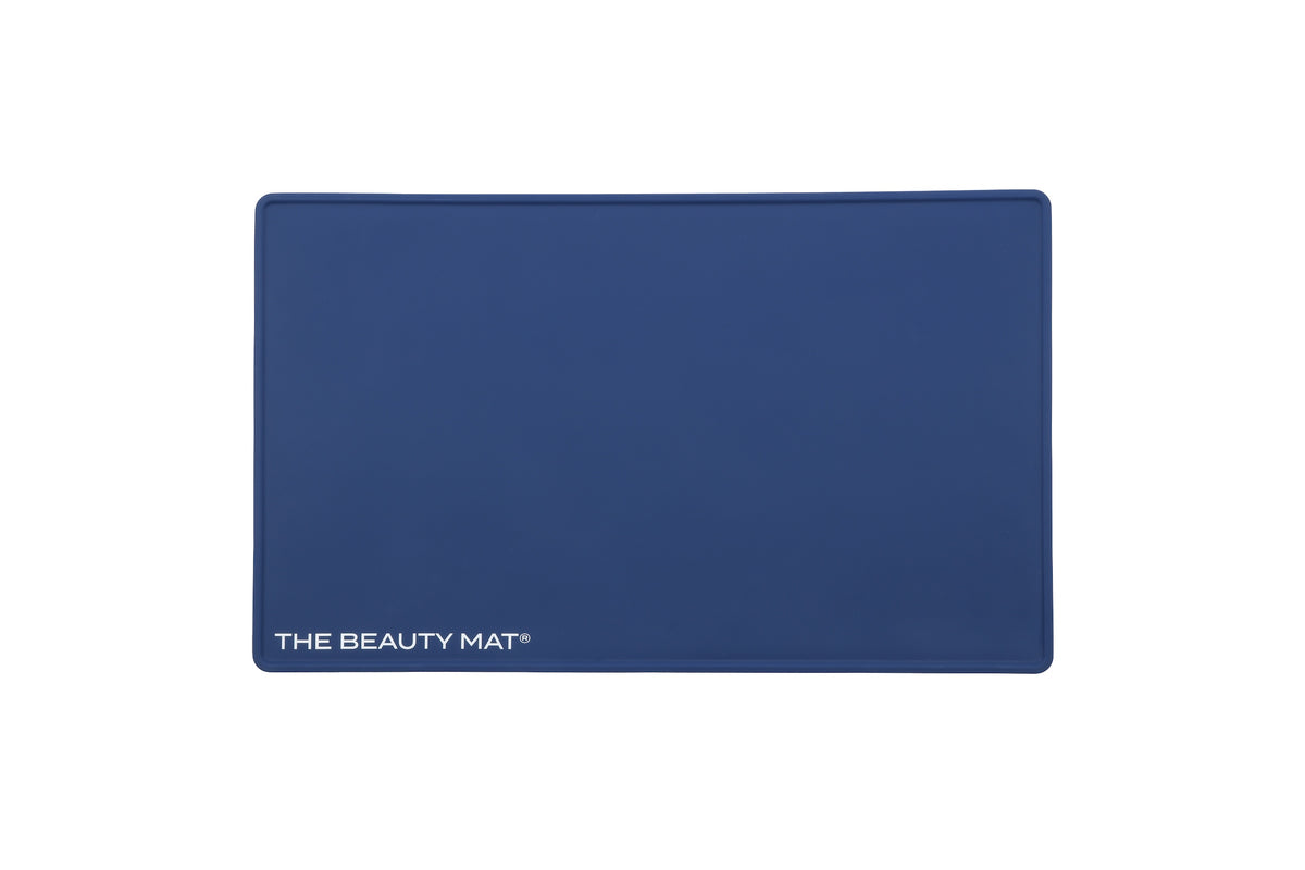 A rectangular navy blue beauty mat with rounded corners and the text 'THE BEAUTY MAT' printed in white at the bottom left, shown on a plain white background.