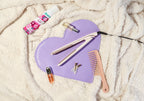 A flat iron hair straightener and styling tools set arranged on a lilac heart-shaped beauty mat, styled on a cream textured blanket with a hair bottle, bobby pins, and a pink comb.