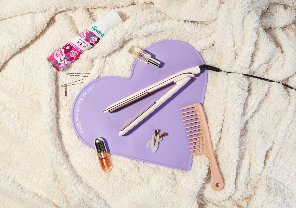 A flat iron hair straightener and styling tools set arranged on a lilac heart-shaped beauty mat, styled on a cream textured blanket with a hair bottle, bobby pins, and a pink comb.