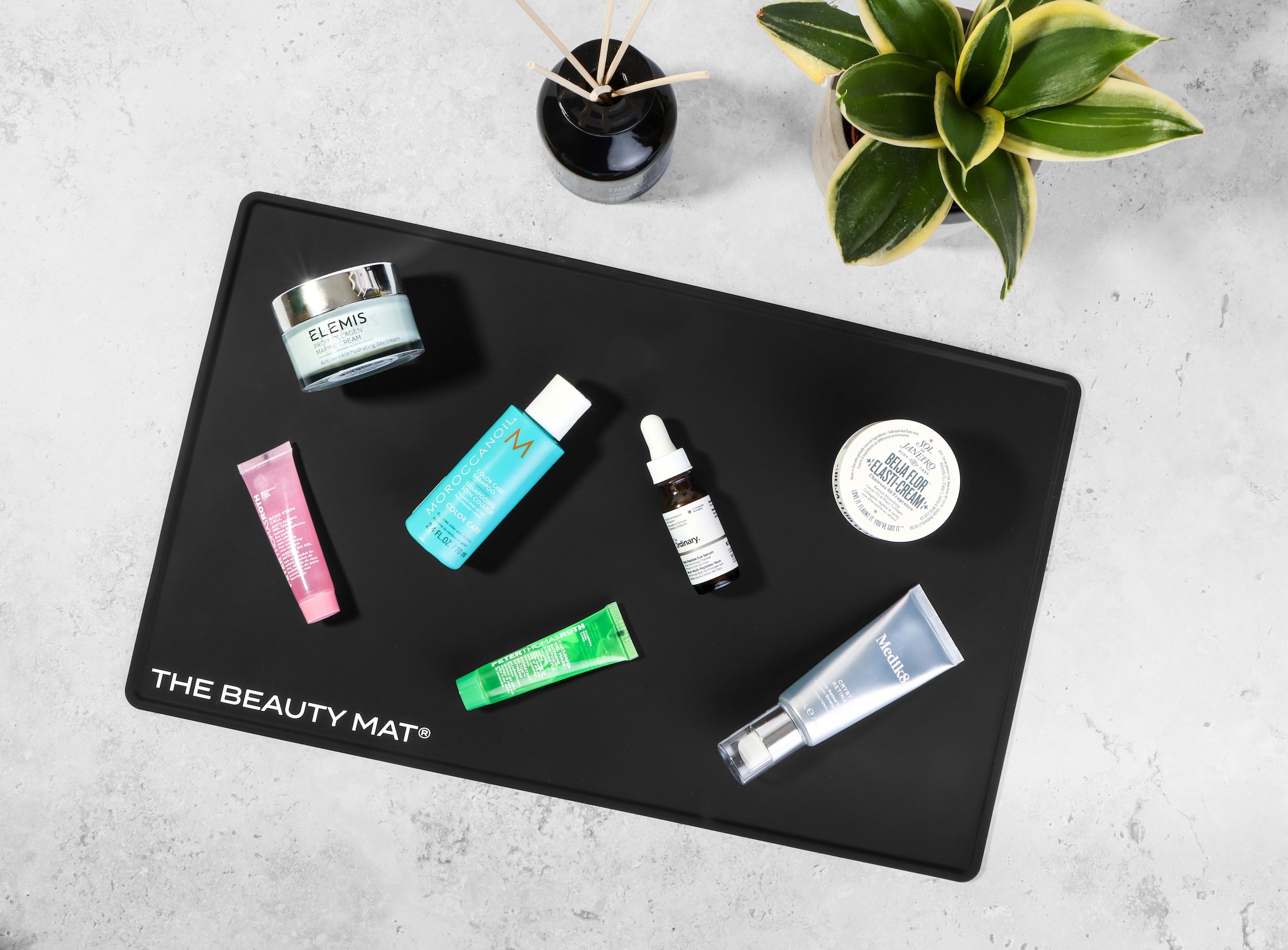 A black rectangular mat labeled 'THE BEAUTY MAT' is arranged on a light gray textured surface with several cosmetic products placed on top, including a pink tube, a turquoise bottle, a green tube, a white dropper bottle, and a silver tube. A small black reed diffuser and a green variegated plant are visible in the background.