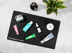 A black rectangular mat labeled 'THE BEAUTY MAT' is arranged on a light gray textured surface with several cosmetic products placed on top, including a pink tube, a turquoise bottle, a green tube, a white dropper bottle, and a silver tube. A small black reed diffuser and a green variegated plant are visible in the background.